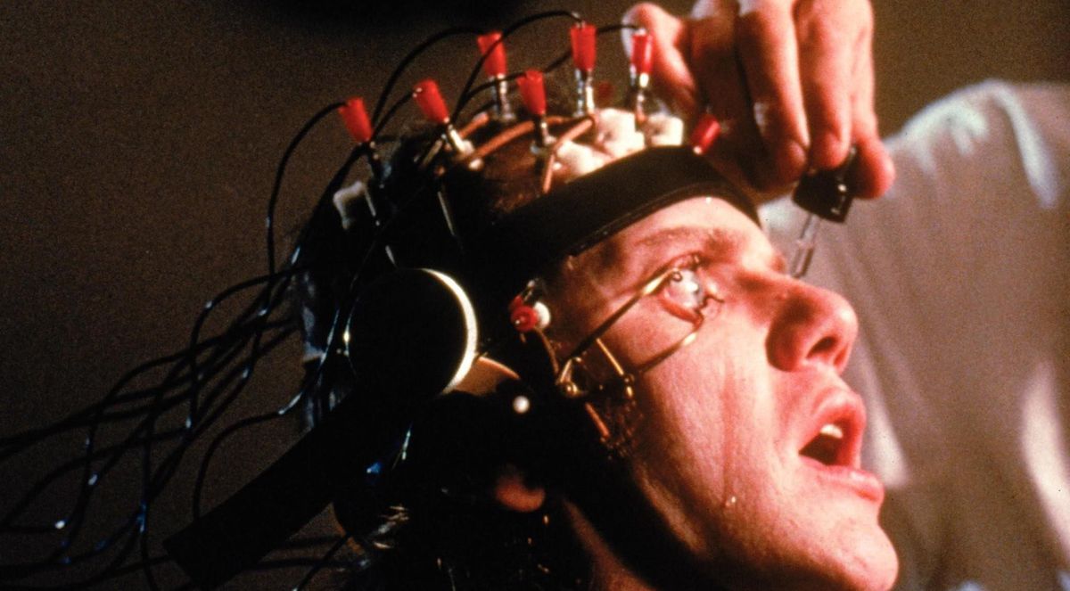 Malcolm McDowell ses i närbild med en metallapparat fastspänd på huvudet och hans ögon hålls öppna med klämmor när en läkare droppar vätska i hans ögon i A Clockwork Orange.