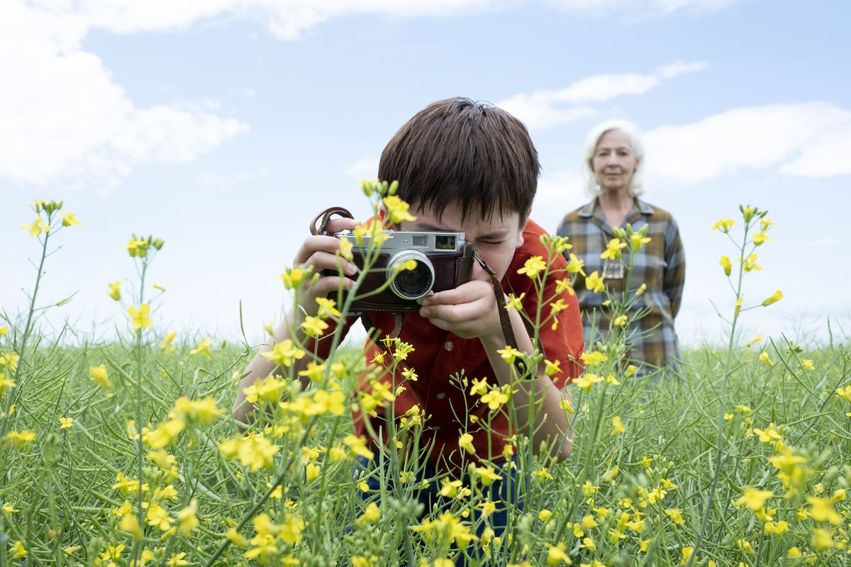 En pojke i en ljusröd skjorta använder en gammaldags manuell kamera för att ta en närbild av en ljusgul blomma när en äldre vithårig kvinna står bakom honom och tittar.