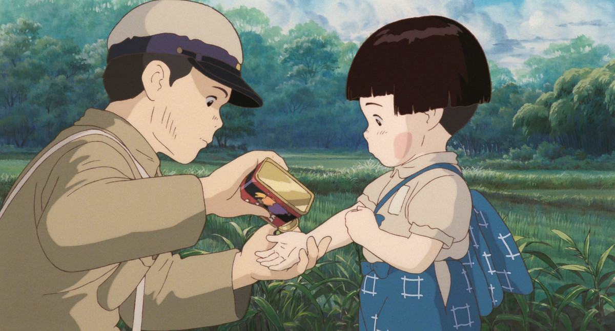 En ung pojke håller sin systers hand när han häller en bit godis ur ett tenn för henne i Grave of the Fireflies