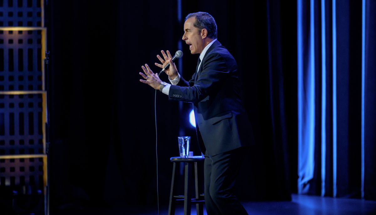 Jerry Seinfeld står på scenen, sett i profil, med händerna spridda ut när han grimaser.