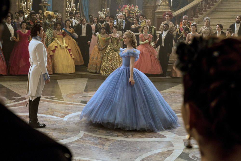 Cinderella (Lily James), in a blue ballgown with an immense skirt and ruffled wraparound neckline, smiles at the prince as the rest of the court stands back and watches in the 2015 live-action Cinderella