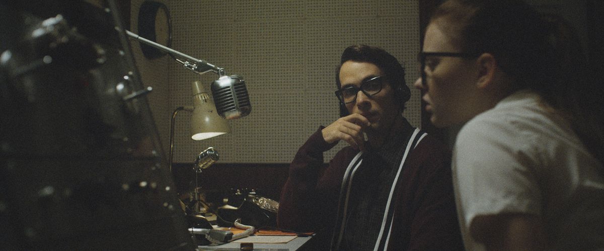 En ung man och kvinna, båda i tunga 1950-glasögon, sitter vid en disk framför en gammal skol tung radiomikrofon i The Vast of Night.