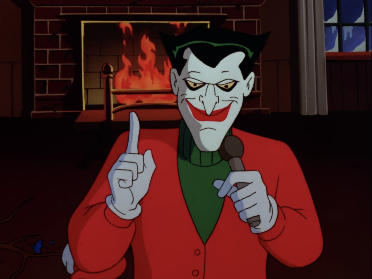 The Joker, dressed for Christmas, addresses the audience in front of a blazing hearth, in “Christmas with the Joker,” from Batman: The Animated Series. 
