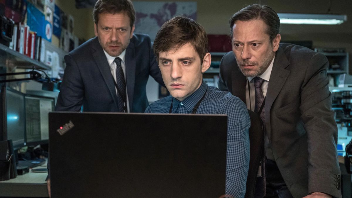 Three men — two wearing suits, one wearing a collared shirt — huddle around an open laptop and stare at it in The Bureau.