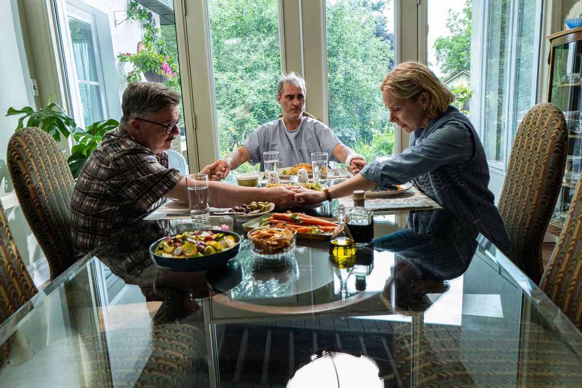 Nathan Lane, Joaquin Phoenix och Amy Ryan sitter runt ett middagsbord och håller varandra i hand och ber i Beau is Afraid