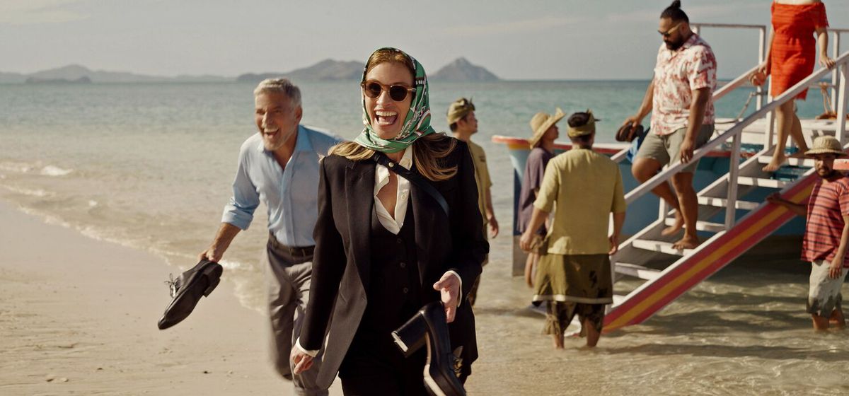En man (George Clooney) och en kvinna som bär en halsduk över huvudet (Julia Roberts) skrattar medan de går av en båt på en strand och håller sina skor i händerna.
