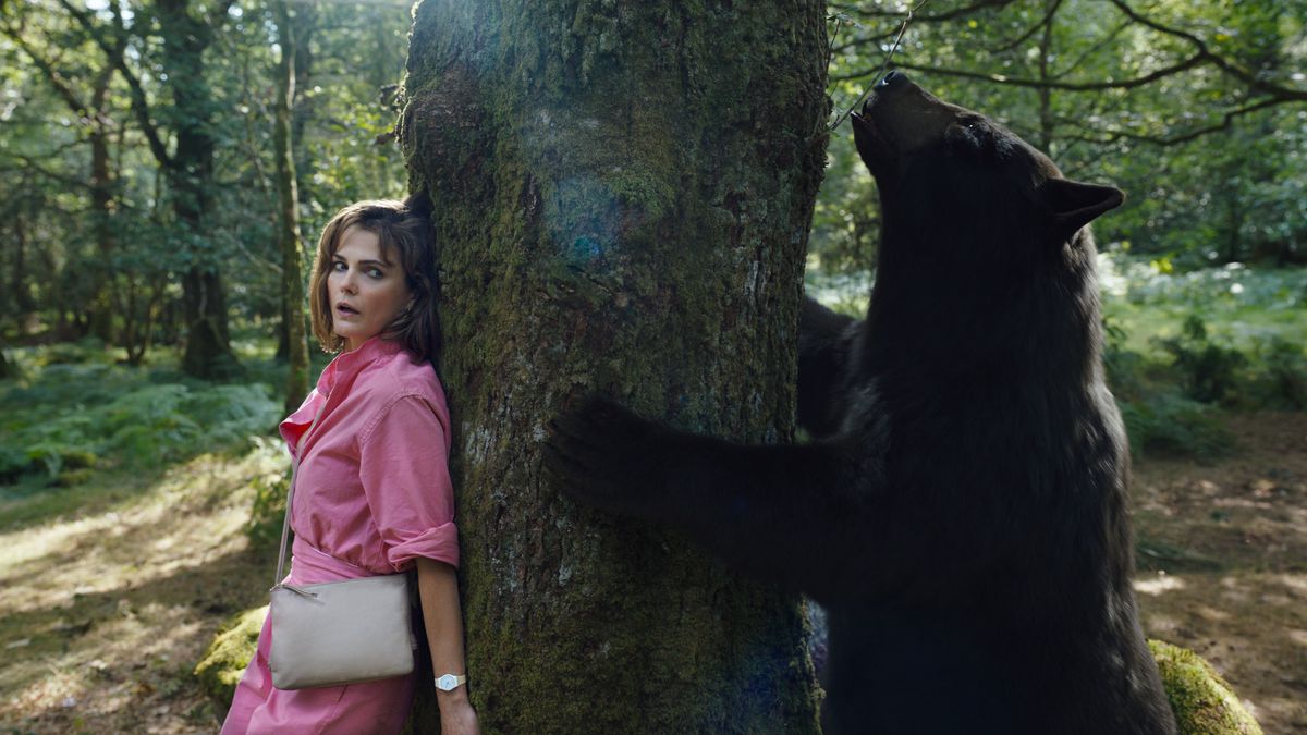 Sari (Keri Russell), en skräckslagen kvinna i en rosa klänning, gömmer sig bakom ett träd när en kokaintillsatt björn snusar runt på andra sidan i Cocaine Bear