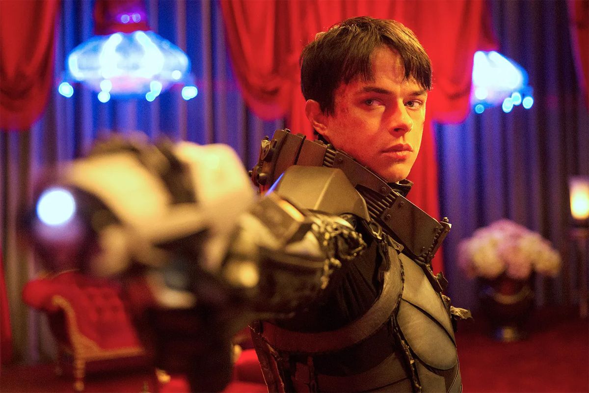 En ung man (Dane DeHaan) med kort svart hår i en högteknologisk rymddräkt siktar in en glödande pistol i ett rött upplyst rum med blå ljuskronor i bakgrunden i Valerian and the City of a Thousand Planets.