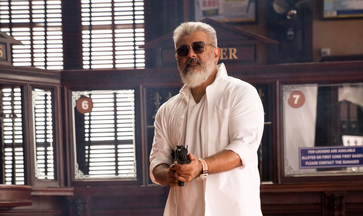 Ajith Kumar holds a sub-machine gun towards the camera while looking cool as hell with all white clothes, white facial hair and hair, and sunglasses. He stands in front of a bank teller booth.