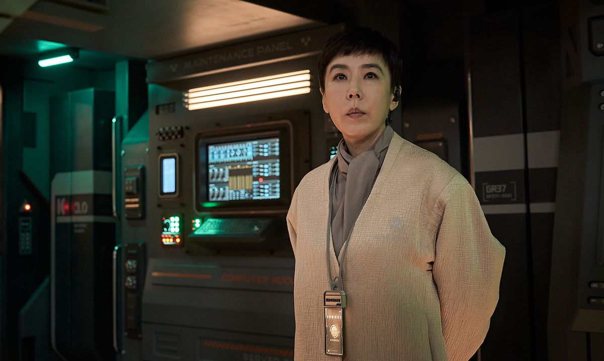A severe-looking woman in a pale linen jacket and futuristic ID tag stands in front of a series of computer displays in Jung_E