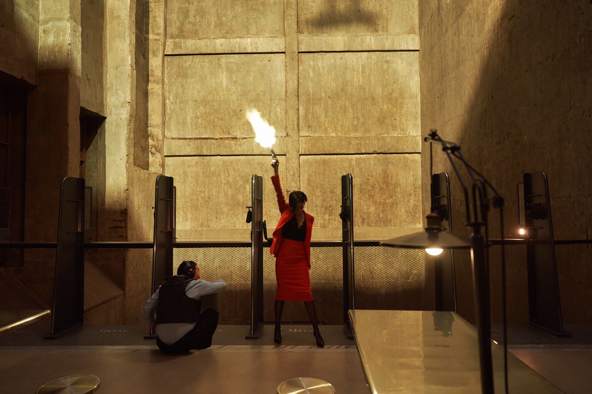 Esom as Cha Min-hee fires a gun in the air while wearing a bright red dress as a police officer crouches down next to her in a firing range in Kill Boksoon.