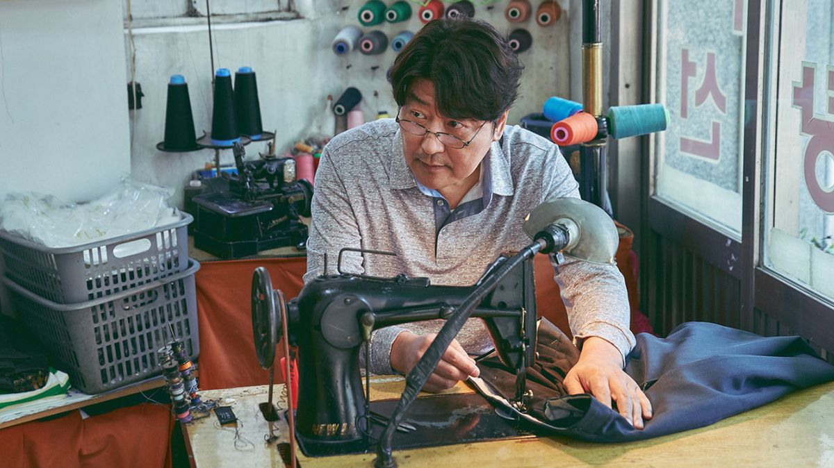 Sang Kang-ho sews some pants while wearing glasses and a collared shirt in Broker. He looks to the right, with an eyebrow raised.