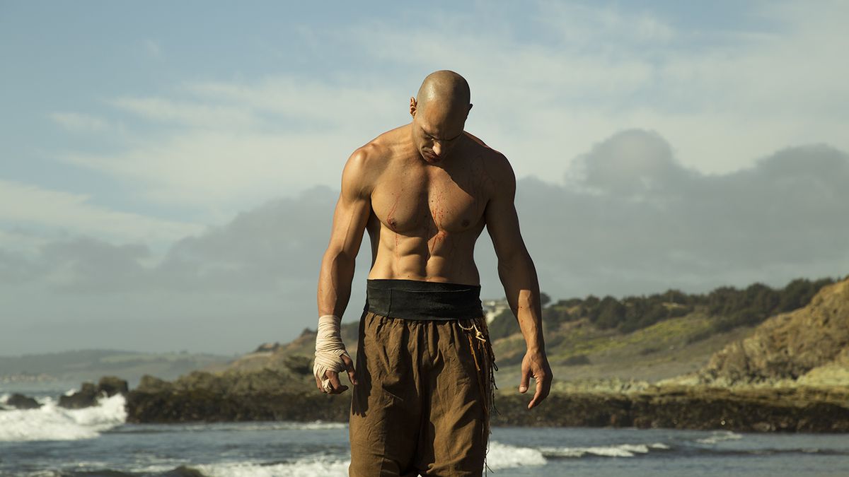 Marko Zaror, shirtless with ripped abs, walks on the beach with his head down in Fist of the Condor.