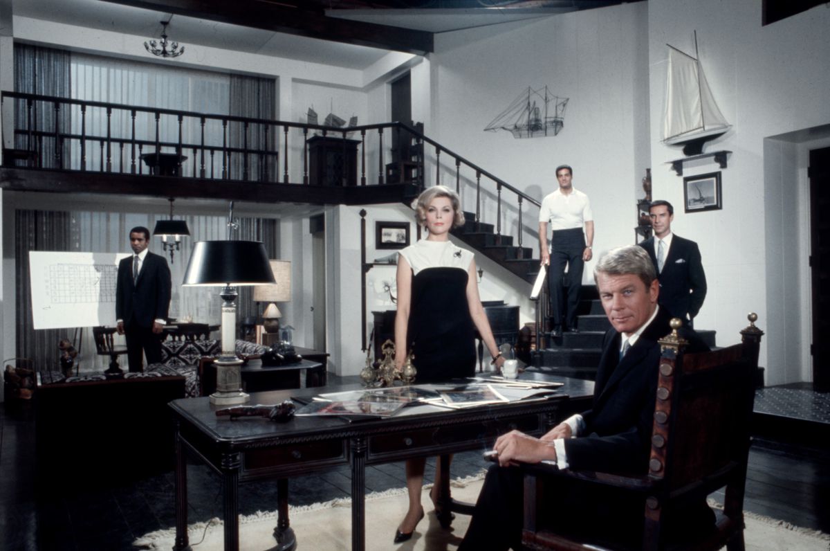 Greg Morris, Barbara Bain, Peter Lupus, Peter Graves, Martin Landau are arranged around a table and stairs in a cast photo of the original Mission: Impossible TV series.