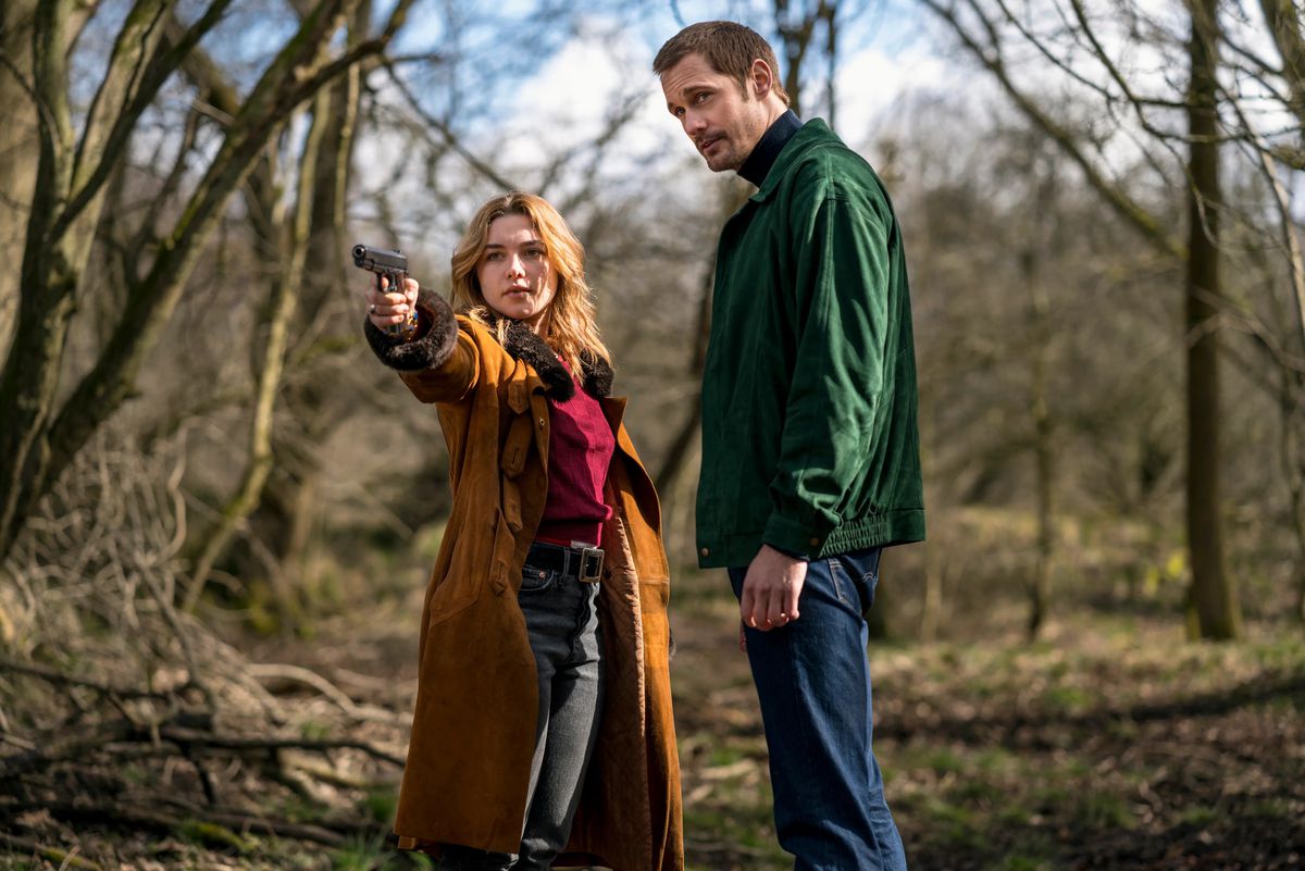 Florence Pugh points a gun while wearing a large orange coat while Alexander Skarsgård stands next to her in a woody area in The Little Drummer Girl.