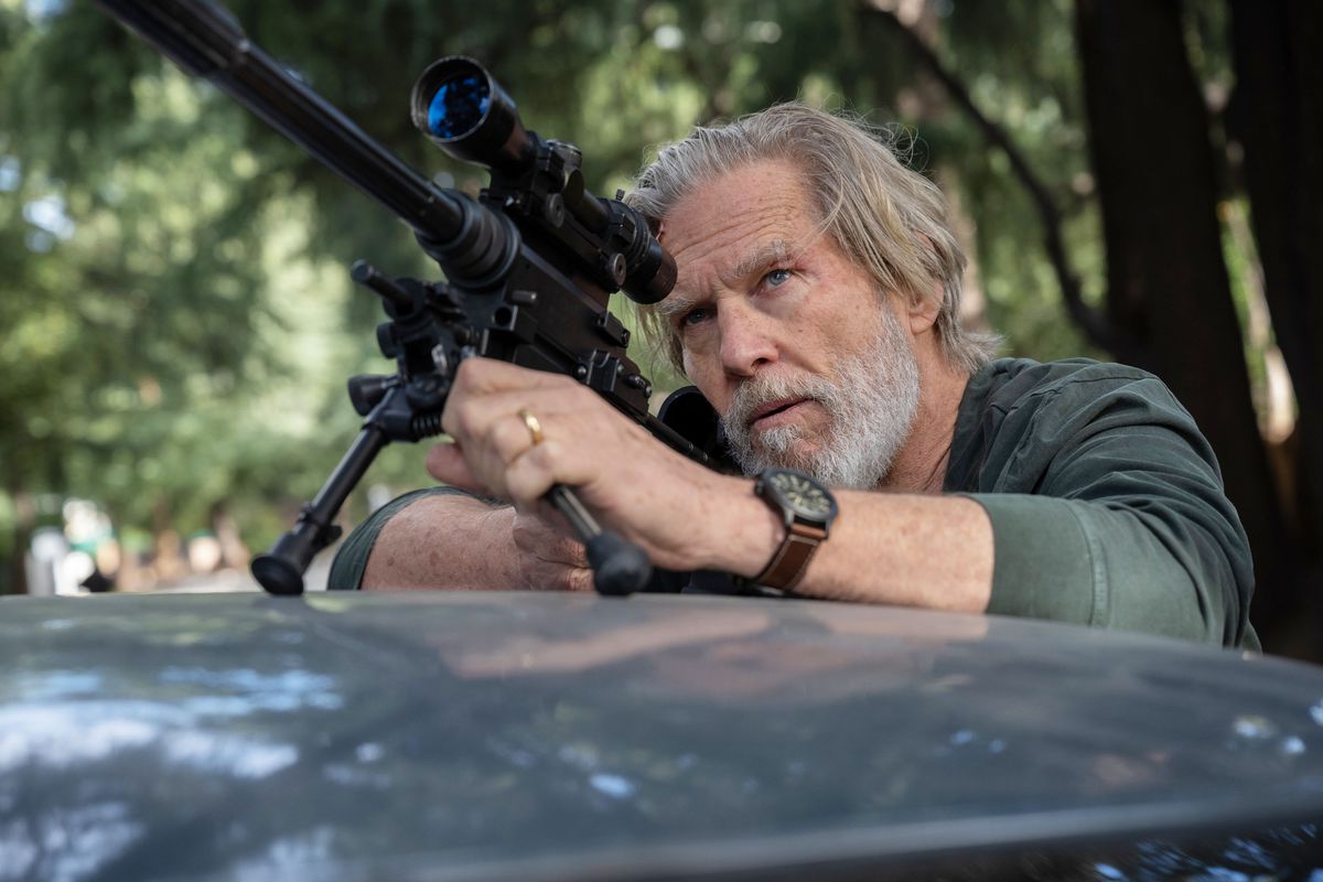 Jeff Bridges, wearing a dark green long sleeve shirt and a watch, peers through a scoped rifle on top of a car in The Old Man.