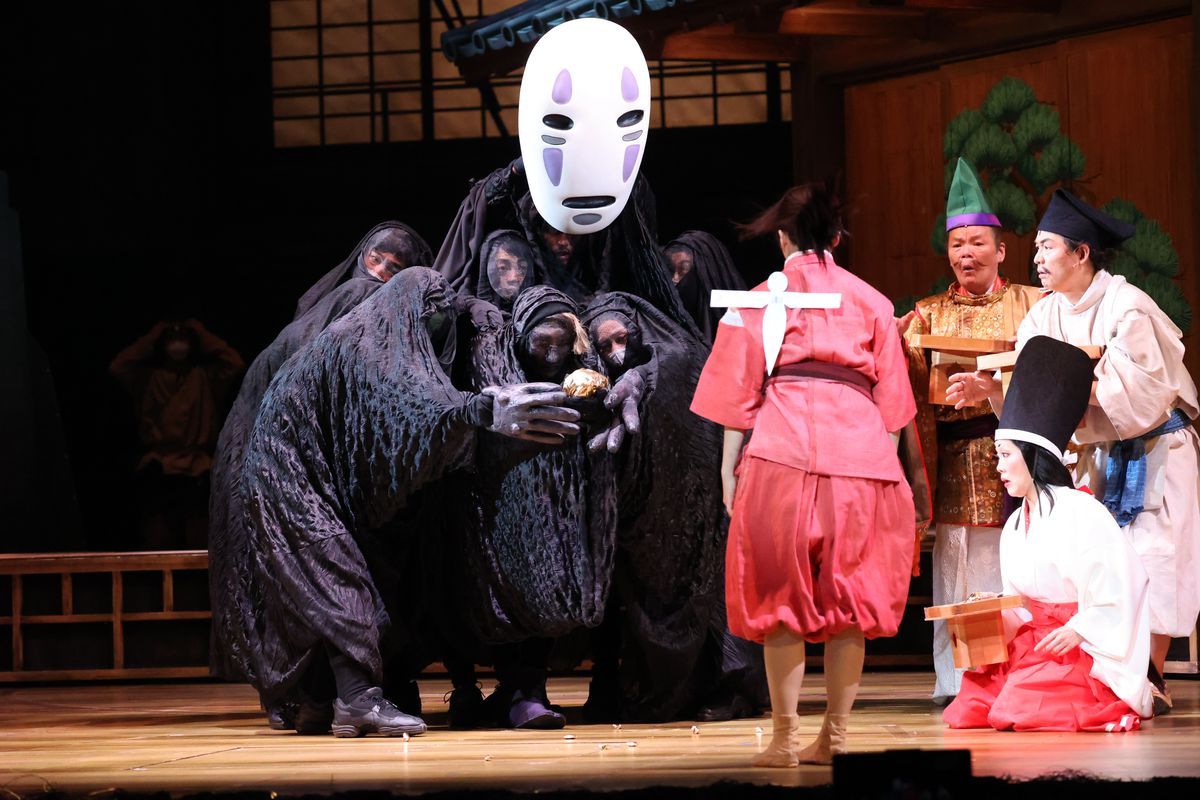 Chihiro (Mone Kamishiraishi, i rosa badhusrockar, med en enda vit Shikigami fast på ryggen) är vänd ner mot Kaonashi (även känd som No-Face, spelad av en grupp artister under en halvtransparent svart presenning) som andra anställda på anda badhus titta på i Spirited Away: Live on Stage