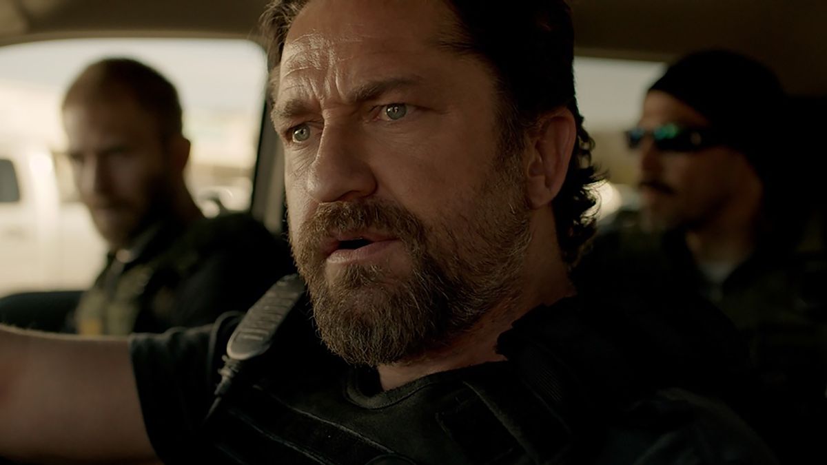 Gerard Butler as “Big Nick” O’Brien in Den of Thieves. He has a goatee and drives a car with two other men in it, all wearing tactical police garb.