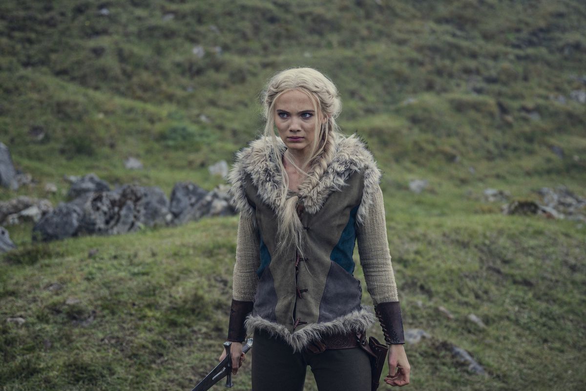 Freya Allen som Ciri i Netflix The Witcher säsong 3 står vänd mot kameran på en gräsbevuxen kulle med ett svärd i handen