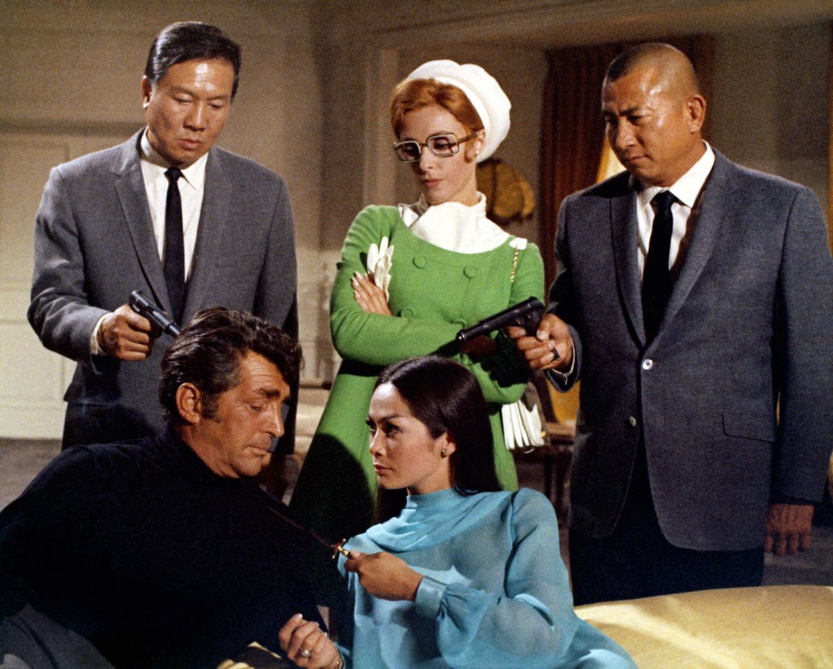 Två asiatiska hantlangare riktar vapen mot Matt Helm när en rödhårig kvinna med glasögon i grön kappa hånar bakom honom och en asiatisk kvinna i blå klänning håller upp en kniv