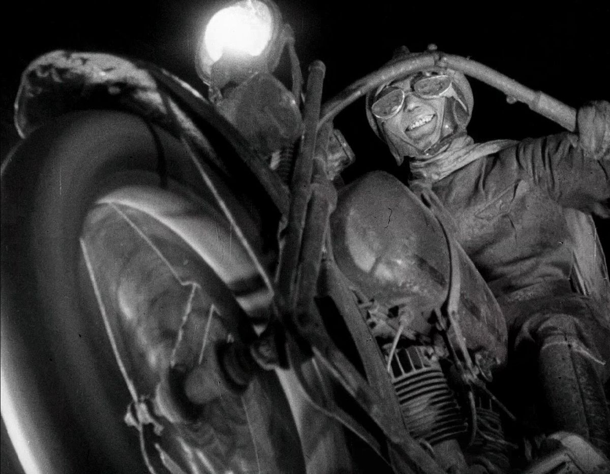 En flinande man i en gammaldags motorcykeldräkt sätter fart på sin motorcykel i Spione