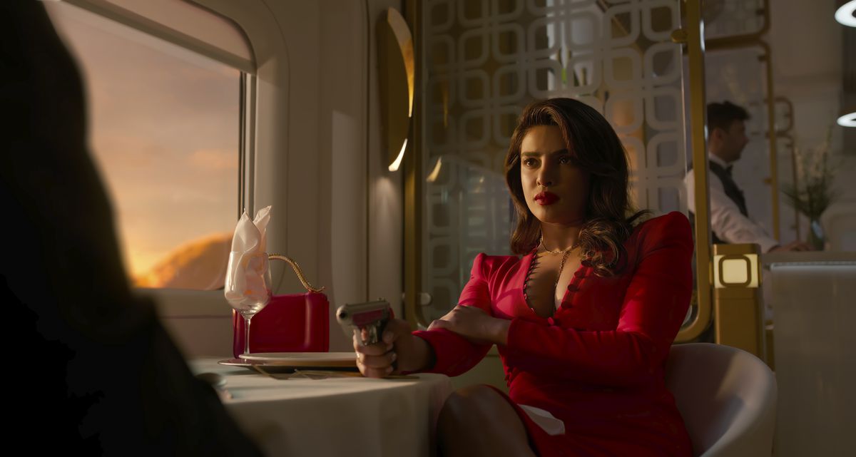 Priyanka Chopra Jonas som Nadia Sinh sitter vid ett matbord på ett tåg när solen går ner genom fönstret och lyser upp hennes röda klänning och pistolen hon pekar på en man utanför skärmen mittemot henne i Prime Video-showen Citadel.