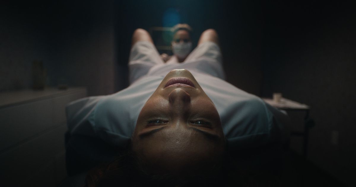 En närbild av en kvinna (Dianna Agron) i en sjukhusklänning som ligger ner på ett bord när en sjuksköterska undersöker mellan hennes ben i Clock.