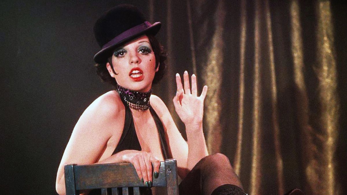 Liza Minnelli wearing makeup, a cabaret costume, and a top hat while performing a dance routine on a chair in Cabaret. 