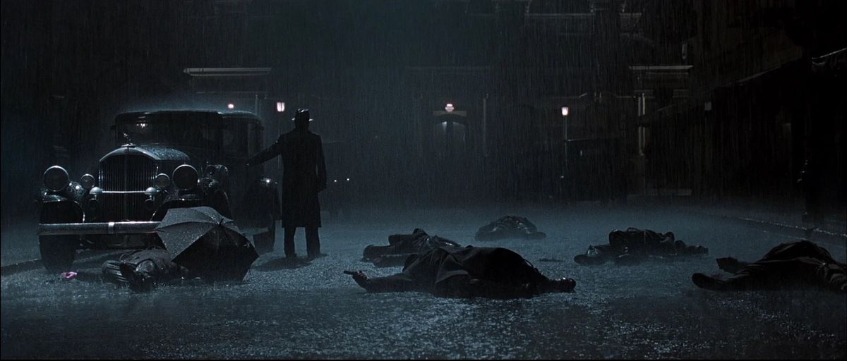 A man in a hat and raincoat stands in the middle of a dark street next to an automobile surrounded by dead bodies as torrential rain pours down.