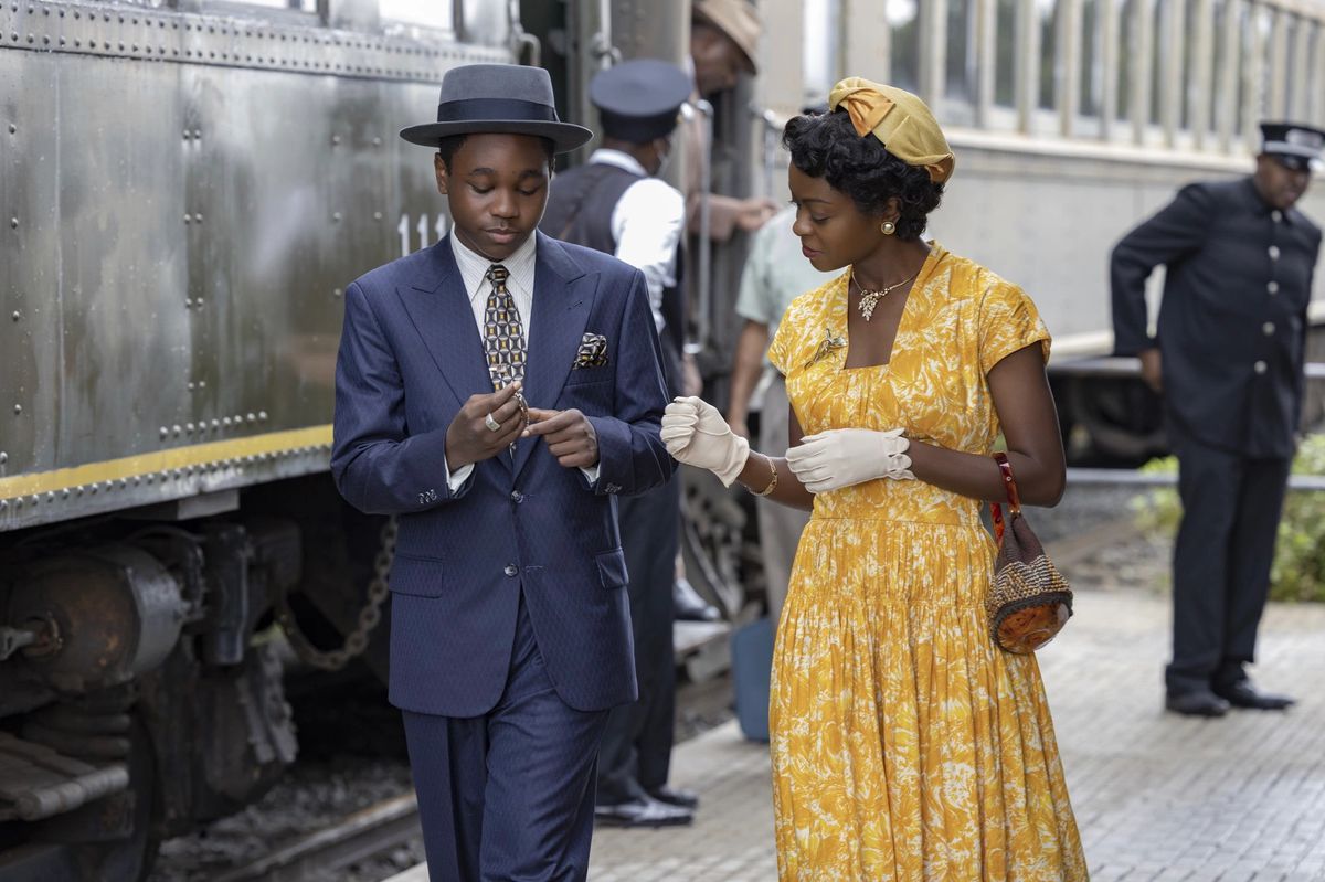 En pojke i en marinblå kostym (Jalyn Hall) står bredvid en kvinna i en gul klänning (Danielle Deadwyler) bredvid ett lokomotiv.