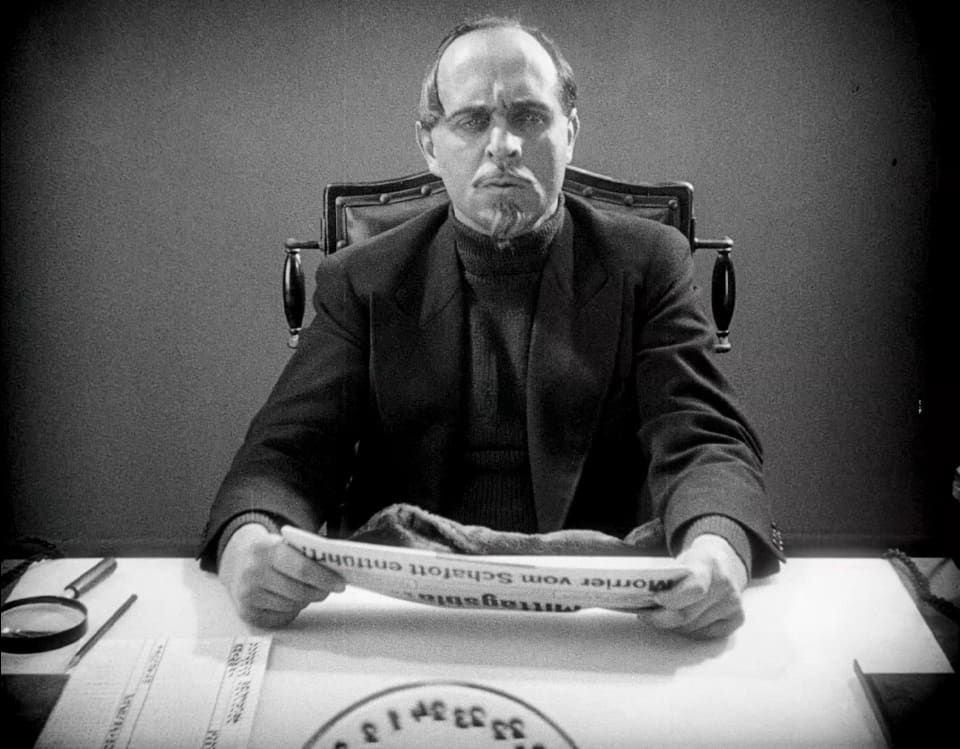 Spione: En man med pärlögda och ett bockskägg sitter i en skrivbordsstol i läder och tittar upp förskräckt på vad han just har läst i en tidning.  Bredvid honom sitter ett förstoringsglas.