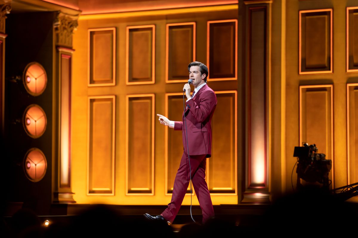 En bred bild av komikern John Mulaney när han går på scenen mot en orange bakgrund i en röd kostym i sin Netflix-special Baby J.