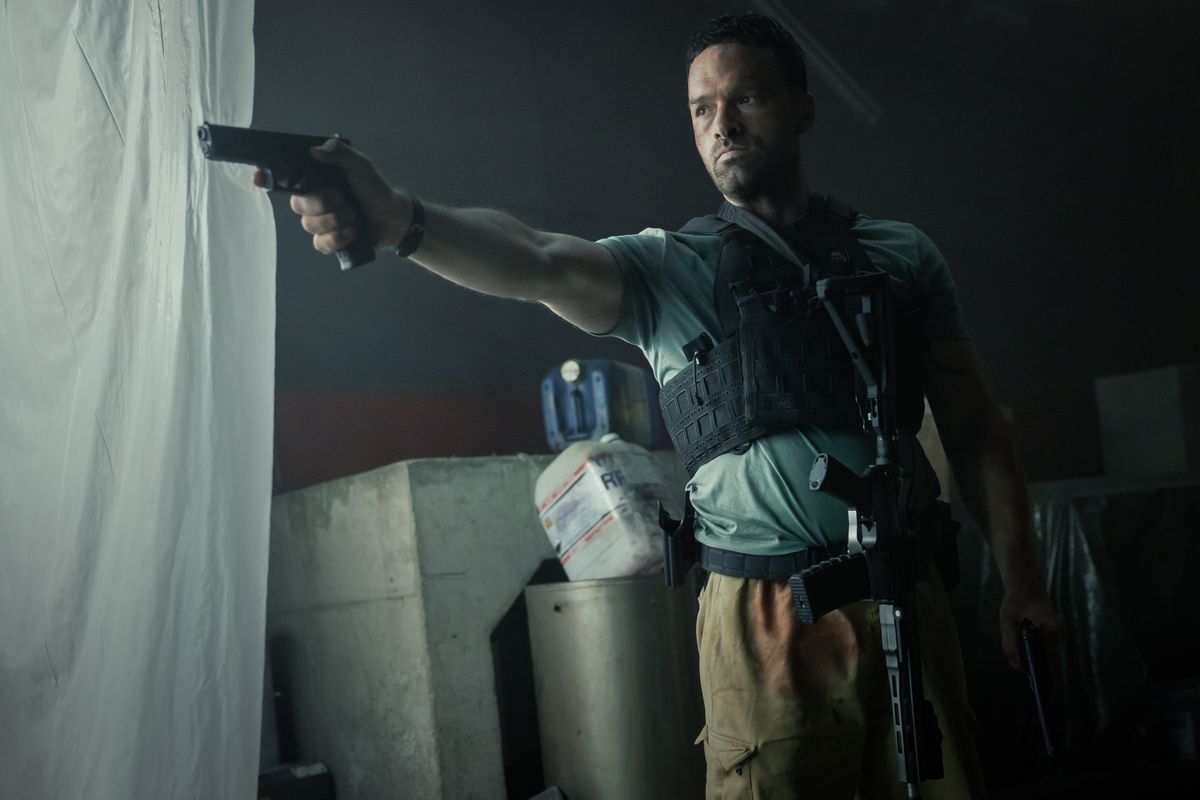 Alban Lenoir som Adam Franco, klädd i en skottsäker väst och ett gevär som hänger i bältet medan han håller en pistol i AKA.