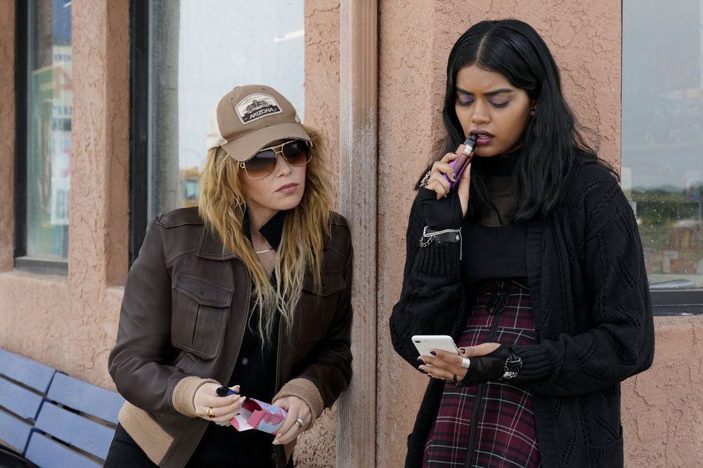 Natasha Lyonne as Charlie Cale, wearing a trucker hat and big sunglasses, leans in as Sara (Megan Suri) nibbles on her vape and looks at her phone in Poker Face