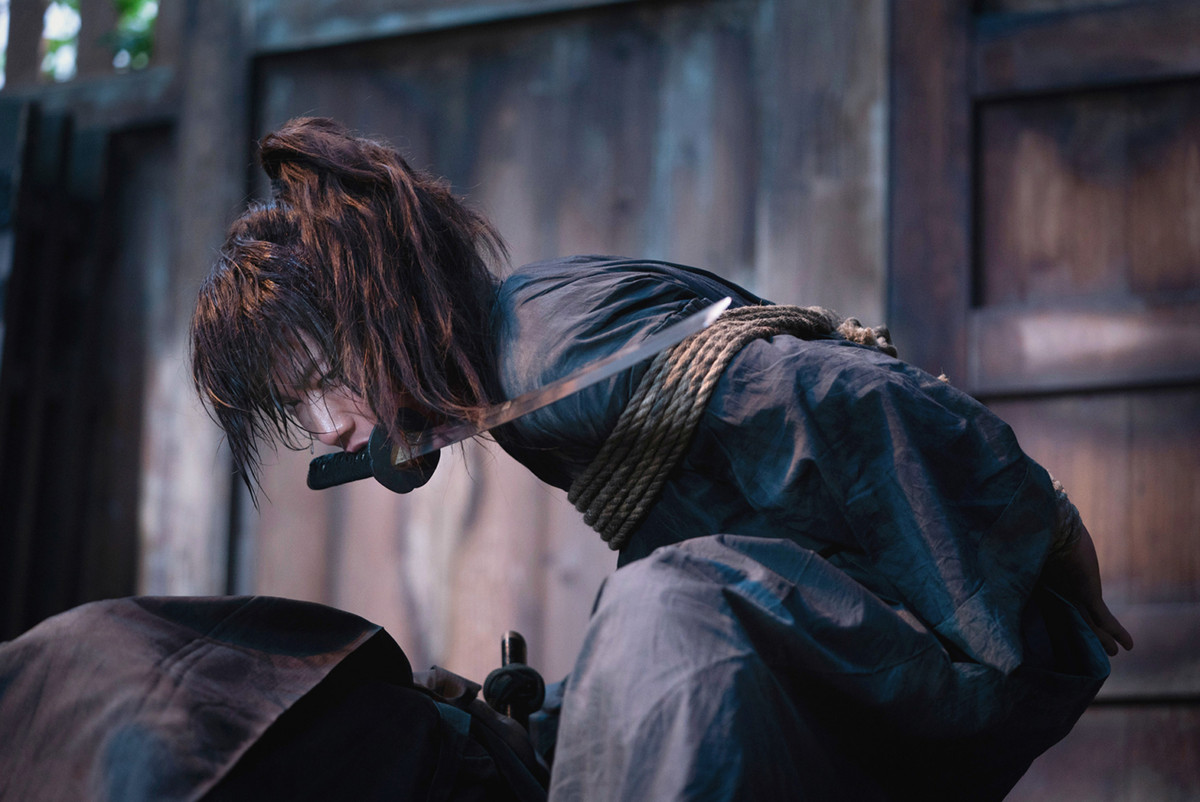 Takeru Satoh as Kenshin Himura holding a sword in his mouth with his hands tied behind his back in Rurouni Kenshin: The Beginning.