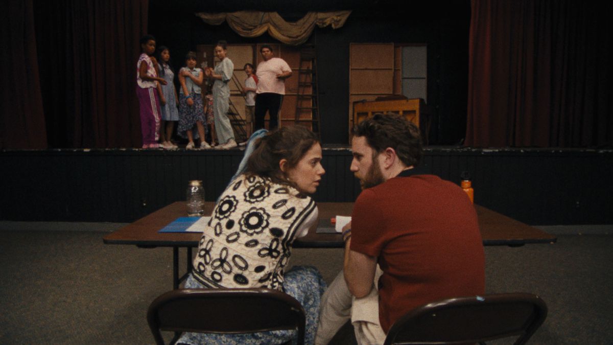 Molly Gordon and Ben Platt talk to each other behind a table in Theater Camp, whilea ctors on stage look on.