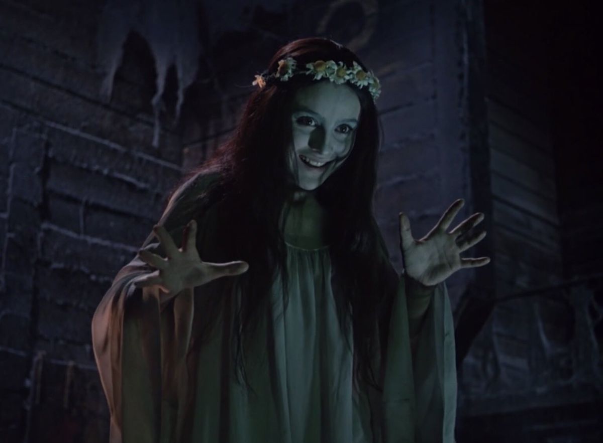 A smiling pale-faced woman wearing a flower crown and a green dress gestures with her hands open in Viy.
