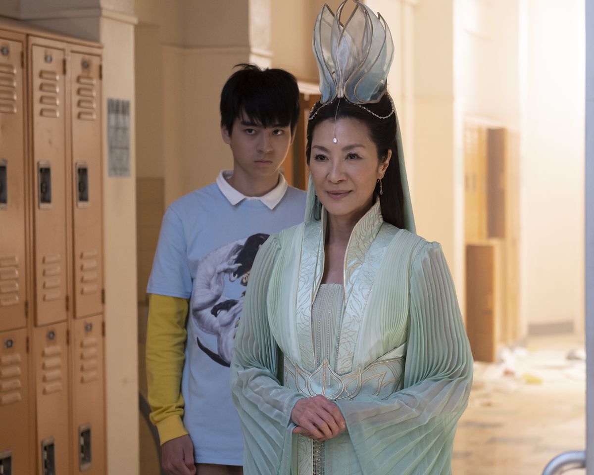 Guanyin (Michelle Yeoh), in fantastical dress, stands smiling in a mundane school hallway lined by lockers. Wei-Chen (Jim Liu) stands behind her frowning in American Born Chinese. 