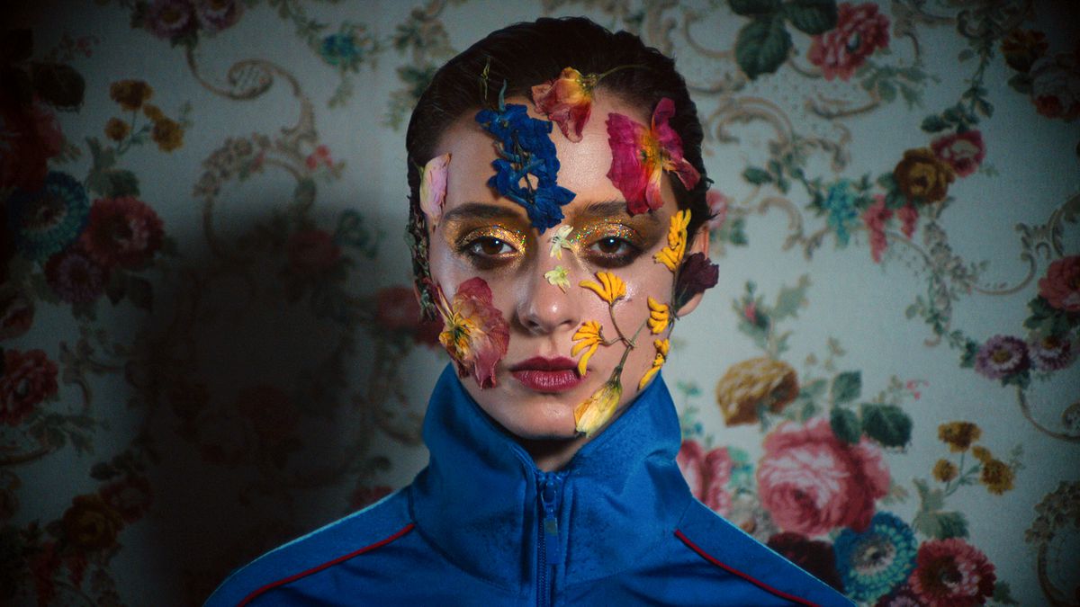 A woman wears a blue jumper in front of a flower-filled wallpaper, while flowers grow out of her face in Copenhagen Cowboy.