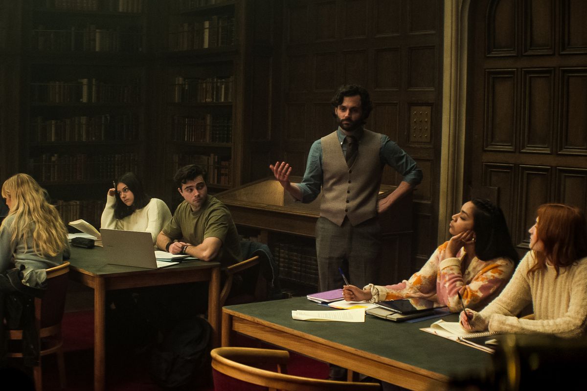 Penn Badgley wears a vest and talks to a group of students in a library in You Season 4.