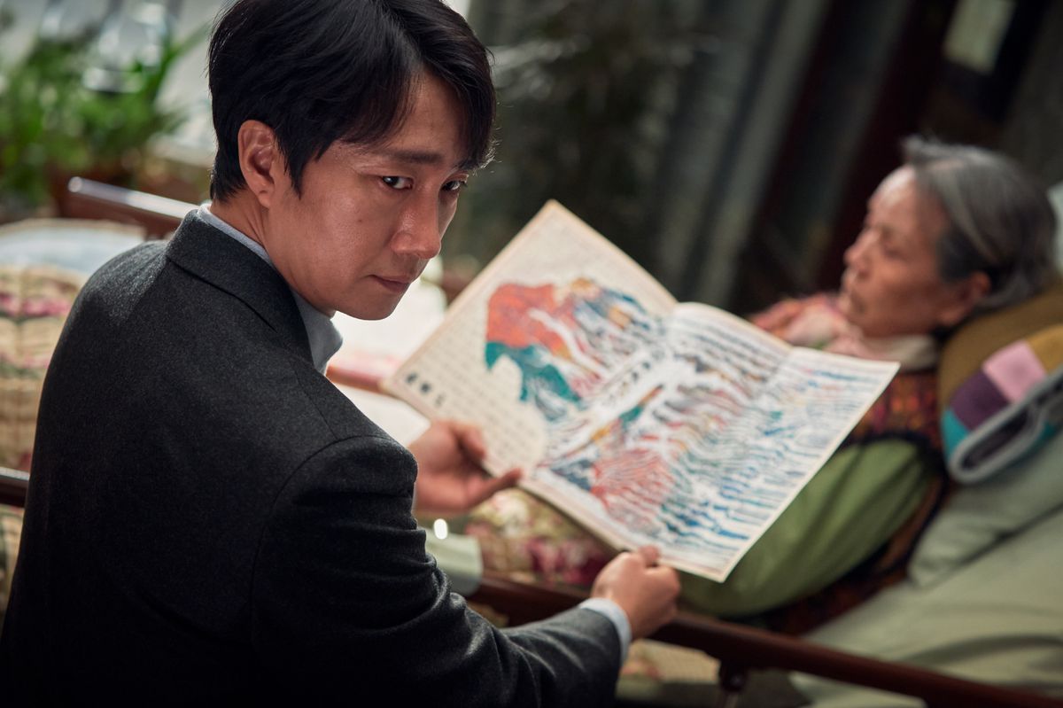 Detective Hae-jun (Park Hae-il) sits next to an old woman and unfolds a colorful map-like page covered with indistinct markings and writing in Decision To Leave