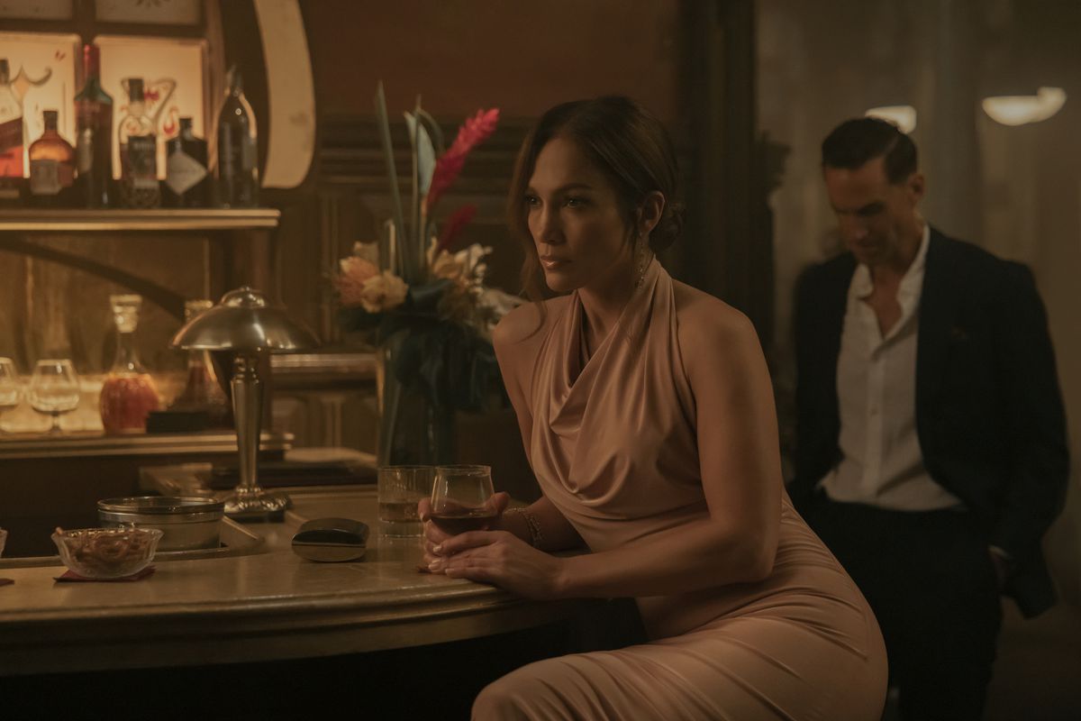 Jennifer Lopez sitter på en bar med ett glas vin i en snygg klänning i The Mother.  Joseph Fiennes skymtar bakom henne, iklädd kostym.