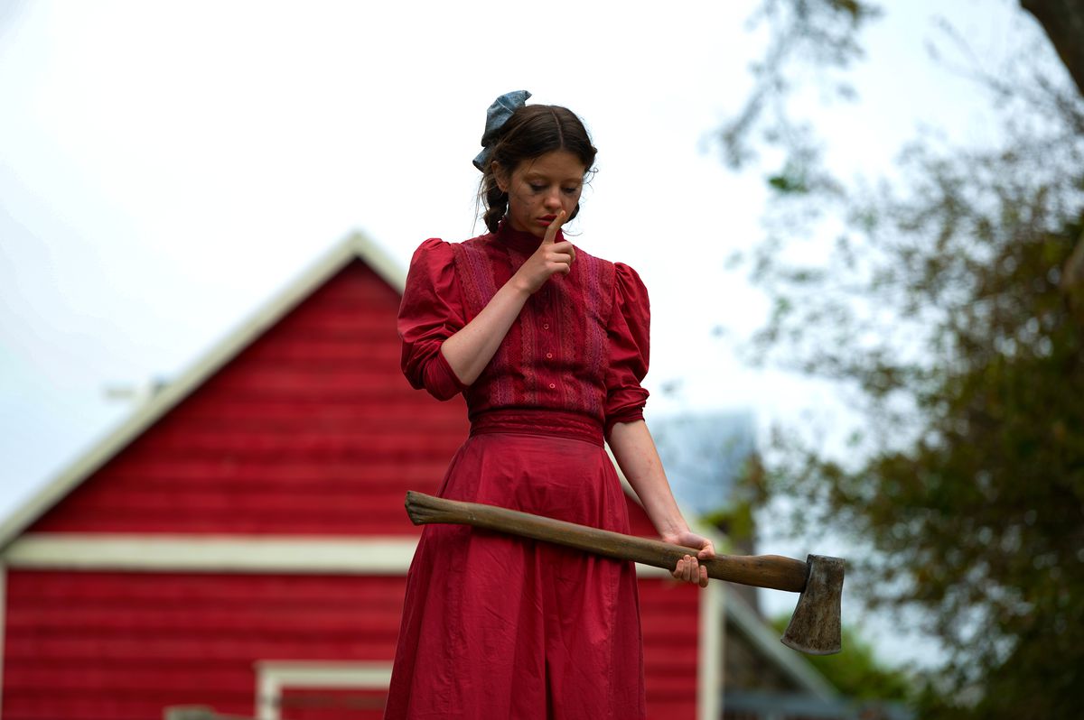 Mia Goth in a vivid red dress, holding an axe and doing a finger-to-the-lips “Shush” in Ti West’s Pearl
