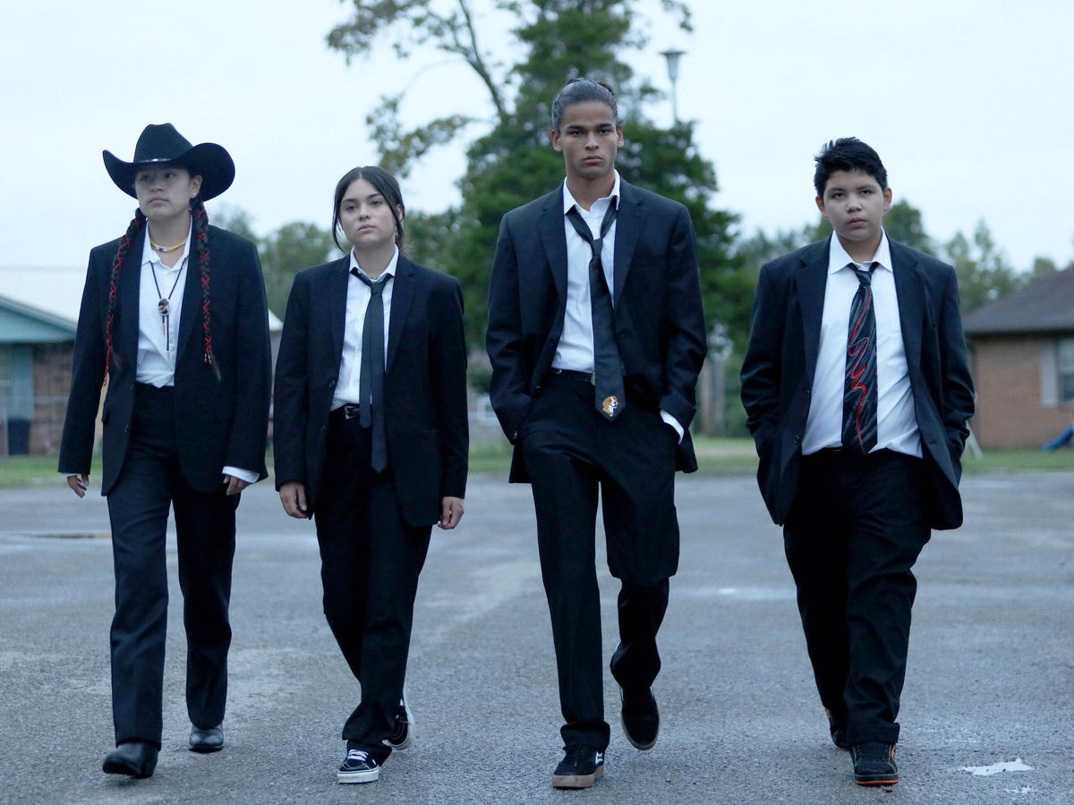 Four teenagers in black suits and ties walking through a parking lot.