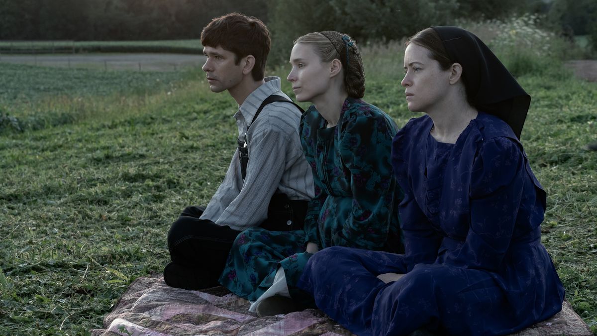En man (Ben Whishaw), en kvinna (Rooney Mara) och en annan kvinna (Claire Foy) sitter på en filt med utsikt över ett fält med grönt gräs.