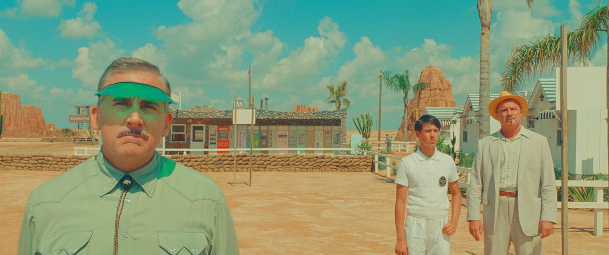 En motellchef (Steve Carell) står framför ett ökenmotell, klädd i ett grönt solskydd och tittar in i kameran, medan två personer i bleka kläder (Aristou Meehan och Liev Schreiber) står på avstånd bakom honom i Wes Andersons Asteroid City
