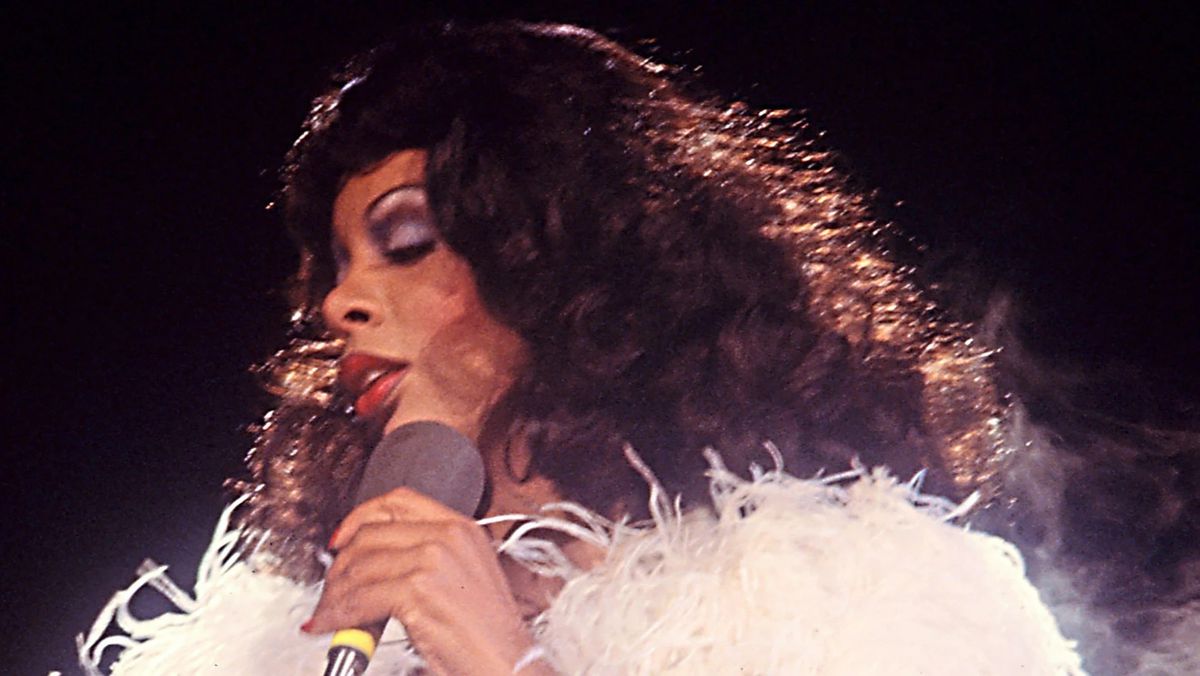 An archival image of Donna Summer in a white feather boa with a microphone in her hand.
