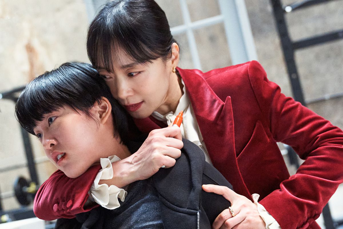 Jeon Do-yeon, wearing a red suit as Gil Boksoon, holds a whiteboard marker to a young girl’s throat in a fight simulation in Kill Boksoon.