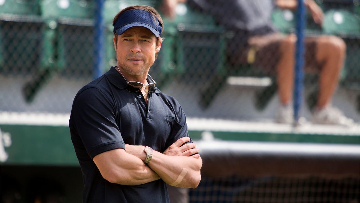 En man i en marinblå pikétröja (Brad Pitt) bär en blå hatt när han står på sidan av en basebollplan.