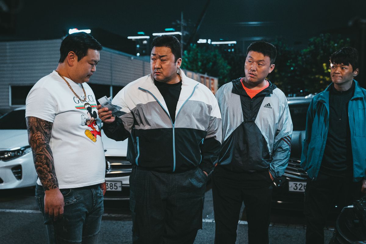 Ma Dong-seok, iklädd träningsoverall, står bredvid en annan kraftig man i en vit t-shirt och två av hans poliskollegor i The Roundup: No Way Out.  Han räcker fram några papperslappar till mannen i den vita t-shirten.